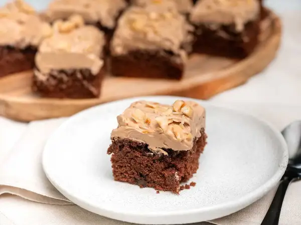 En bit chokladtårta med krämig frosting och nötter. Flera bitar serveras på ett träfat i bakgrunden.