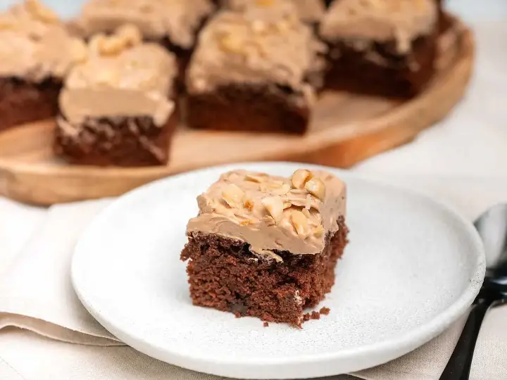 En bit chokladtårta med krämig frosting och nötter. Flera bitar serveras på ett träfat i bakgrunden.